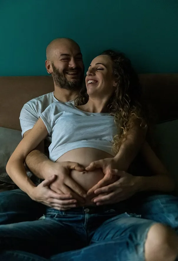 Newborn and fertility photography: expecting couple laughing together with hands forming a heart over the baby bump