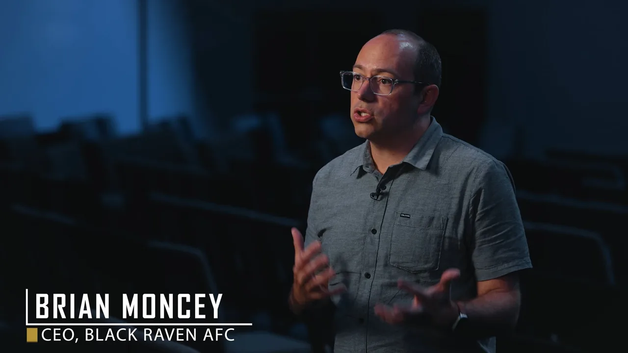 Black Raven AFC case study video: CEO Brian Moncey in glasses and a gray shirt interviewed in a darkened auditorium